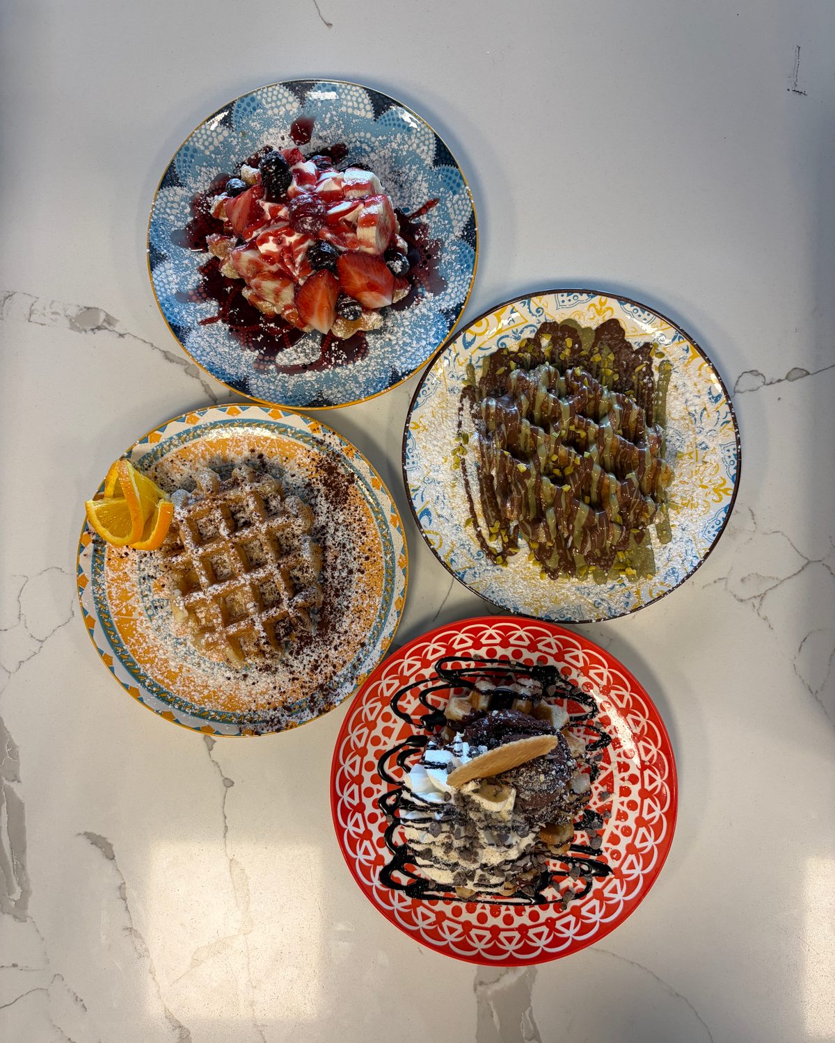 Vier bunte Teller von oben: Waffel mit Orange, Pistazienwaffel, Beerendessert, Schokodessert