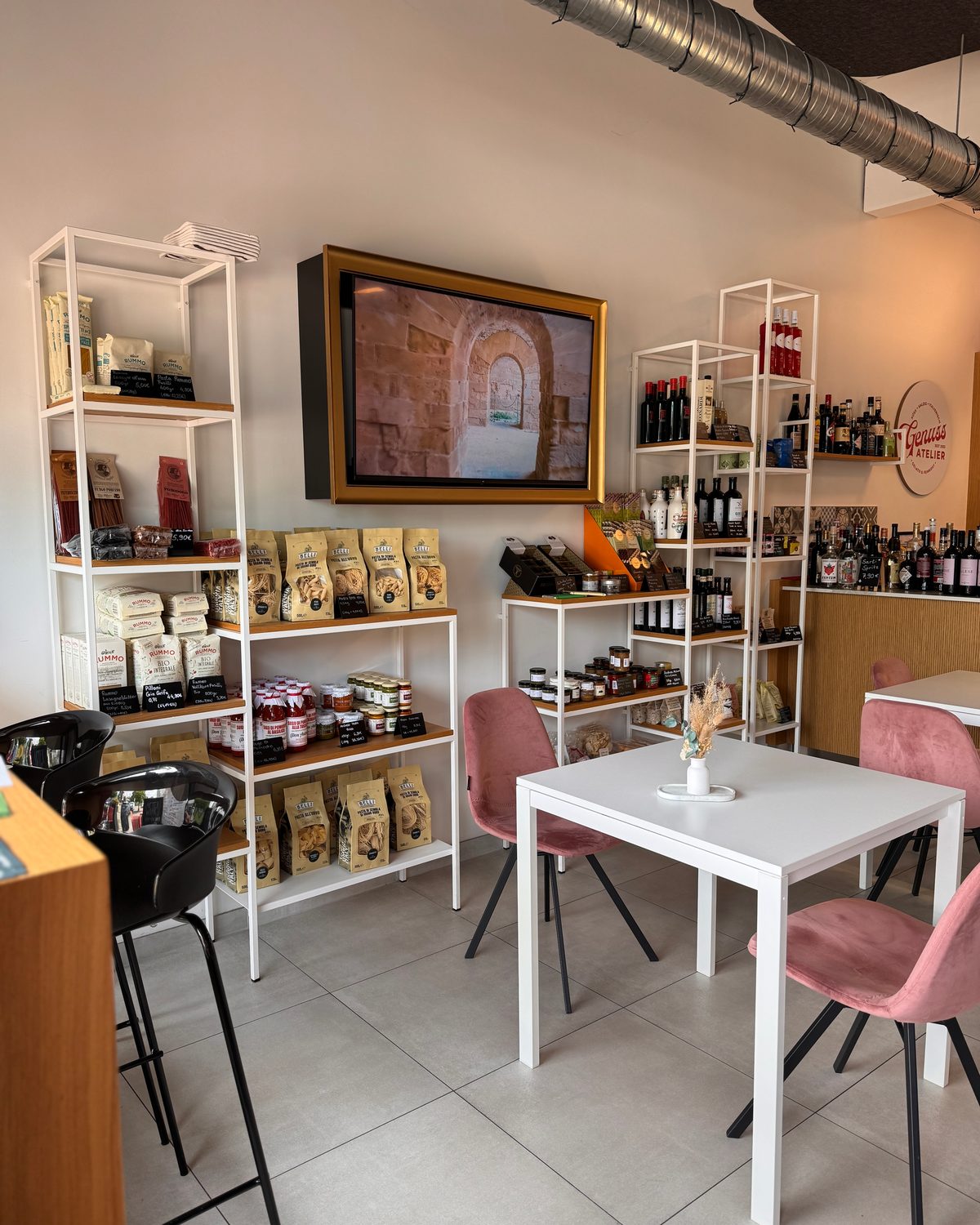 Blick in den Laden: Feinkostregale, Weine, weißer Tisch mit rosa Stühlen und das Genuss-Atelier-Schild