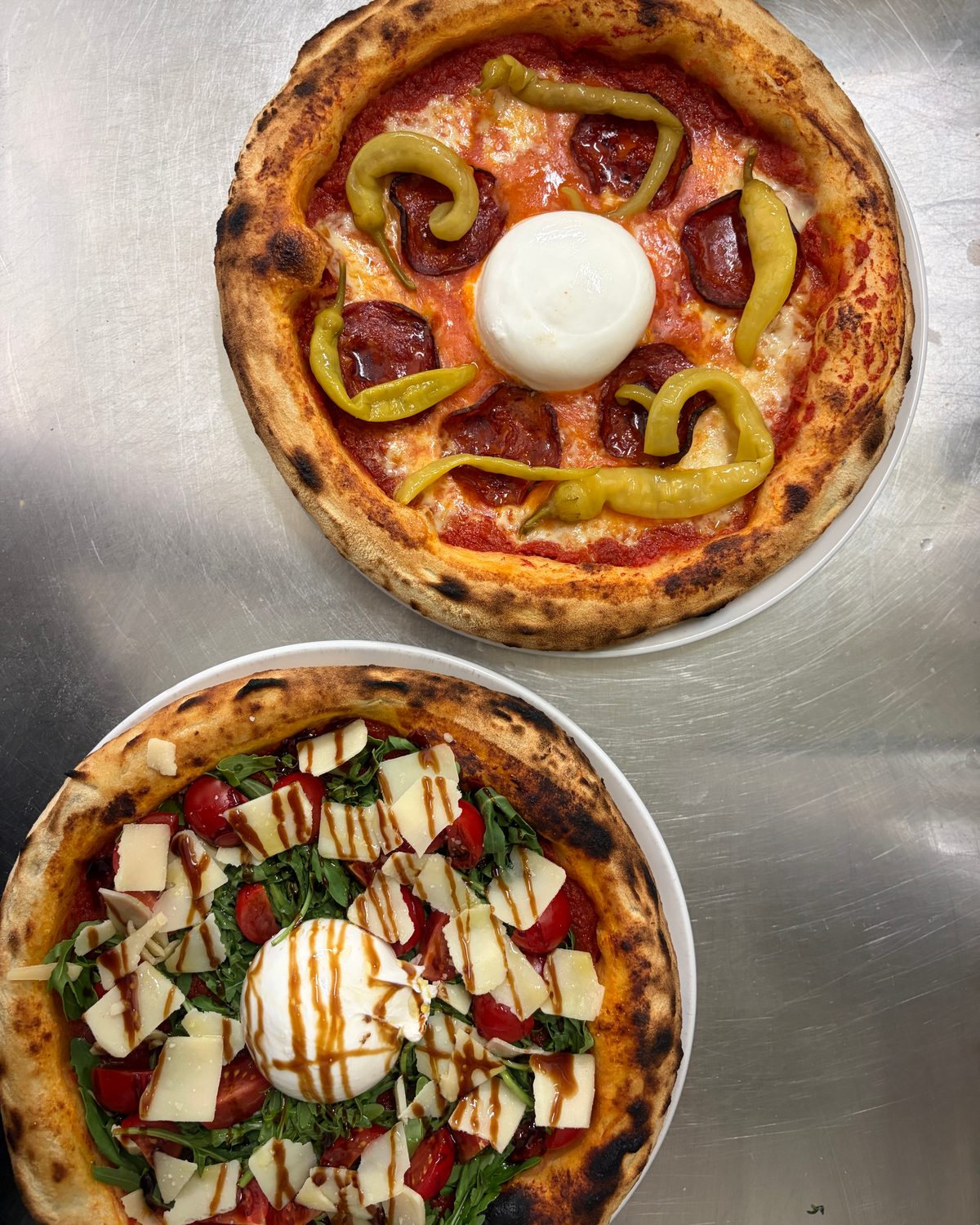 Zwei Pizzen von oben auf Edelstahl: Peperoni mit Burrata, Halloumi mit Rucola