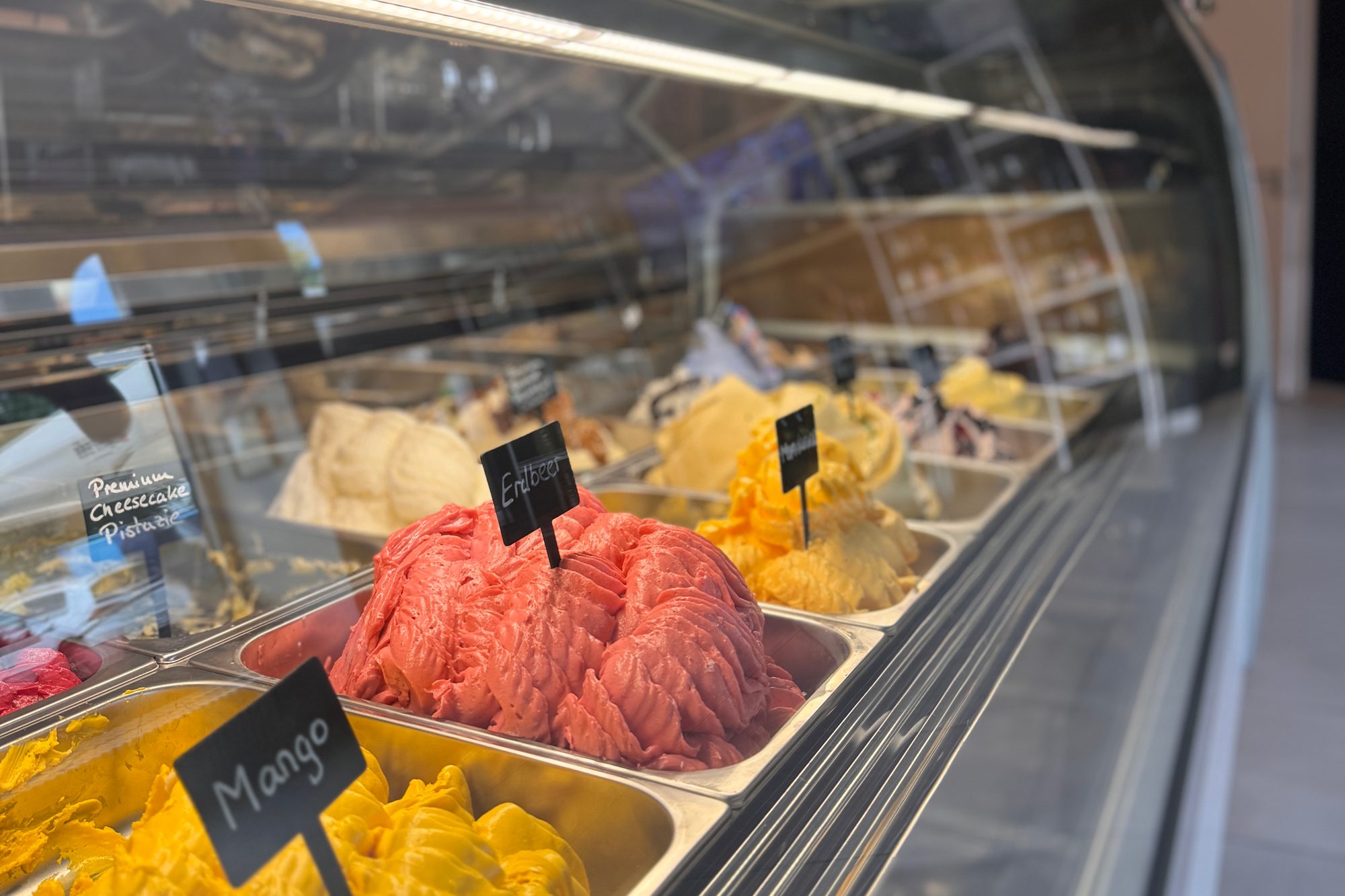 Gelato-Vitrine im Genuss Atelier: lange Glastheke mit frischen Eissorten in Edelstahlwannen, Schildchen mit Sortennamen