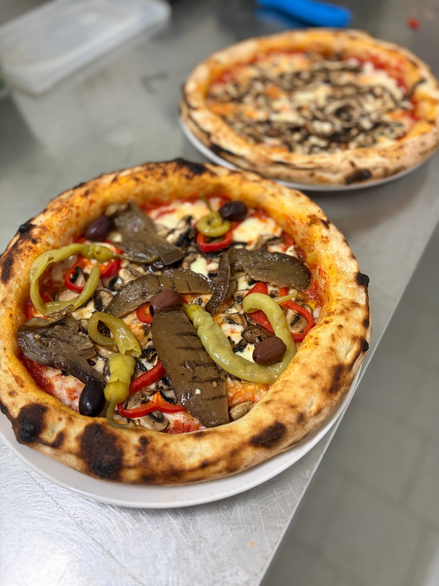 Pizza mit Peperoni, Paprika und Fleisch auf Edelstahl, dahinter eine zweite