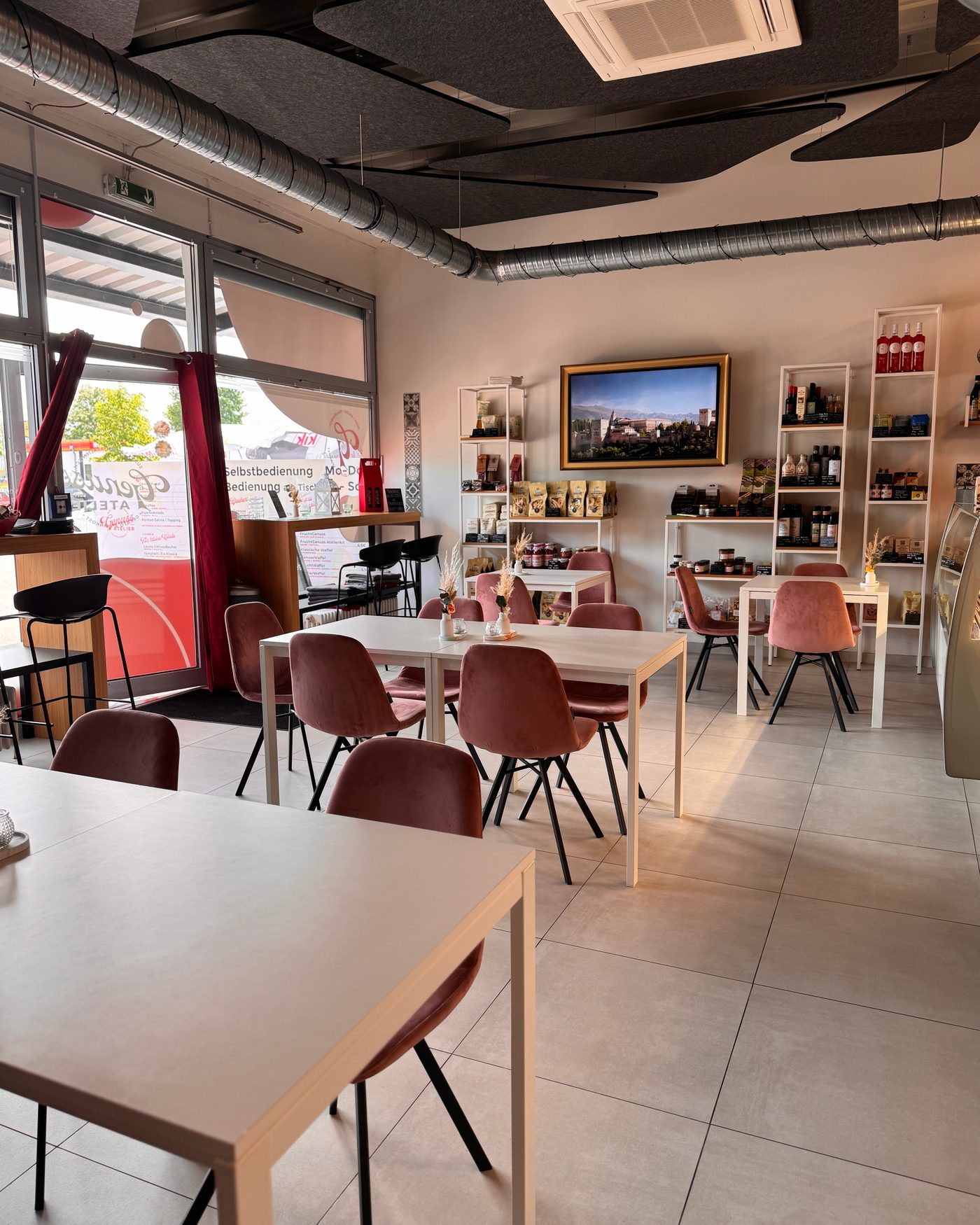 Gastraum des Genuss Atelier: helle Tische, rosa Stühle, Feinkostregale und Bild von Italien an der Wand