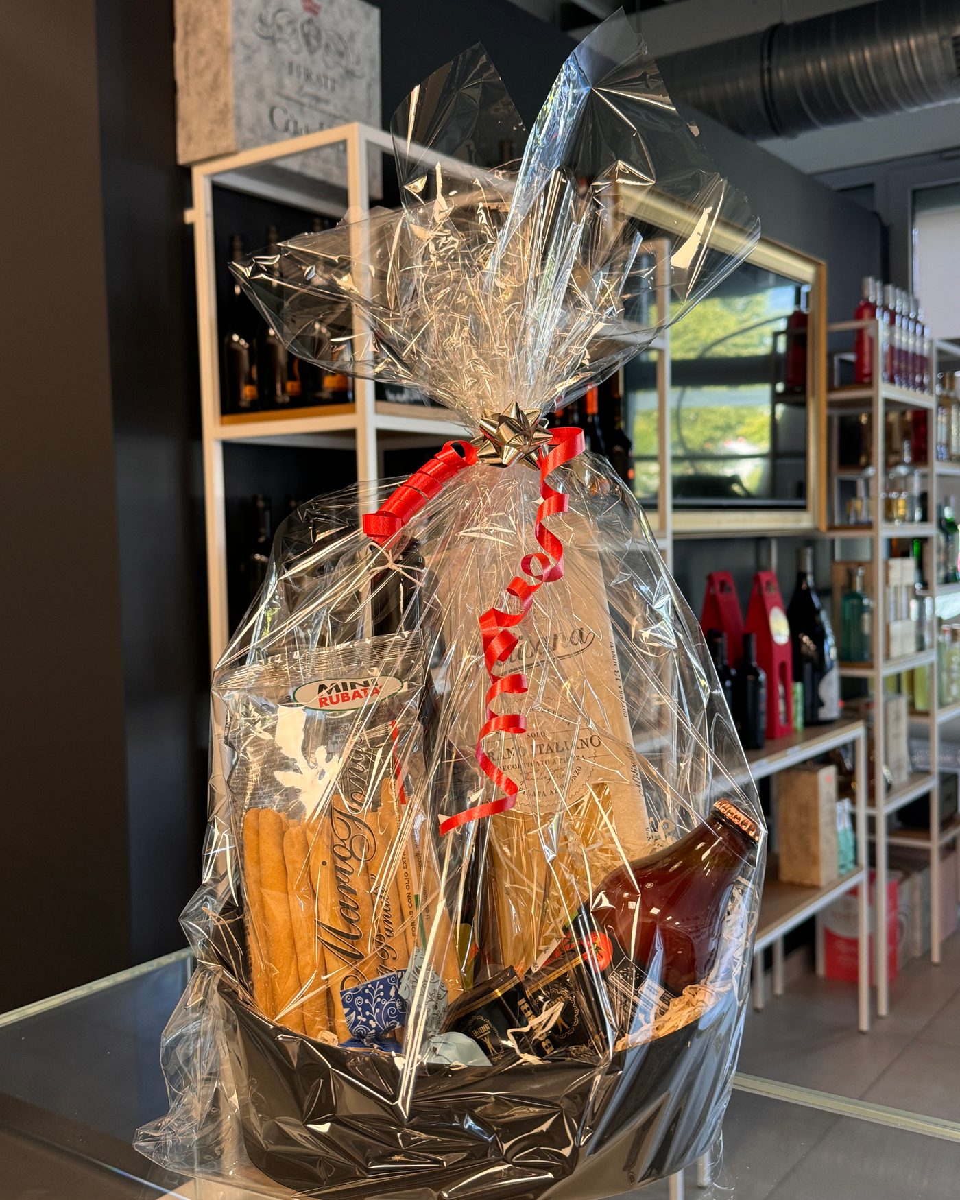 Präsentkorb in Folie mit roter Schleife, gefüllt mit italienischer Pasta, Sugo und Feinkost, vor den Laden-Regalen