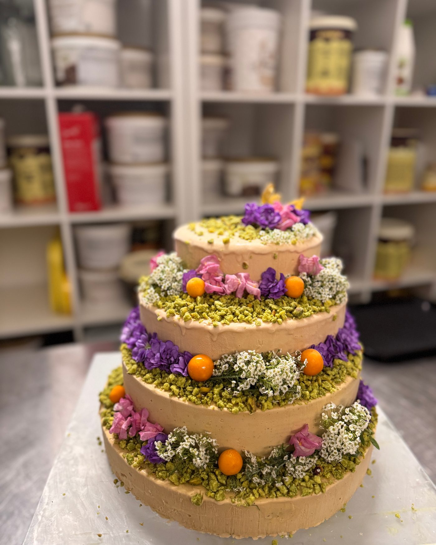 Vierstöckige Torte mit Pistazienrand, lila und weißen Blüten und kleinen Orangen, in der Backstube